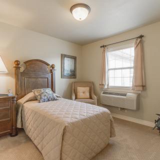 A cozy bedroom with a single bed, dressed in beige tones, featuring an armchair and a side table with a lamp.