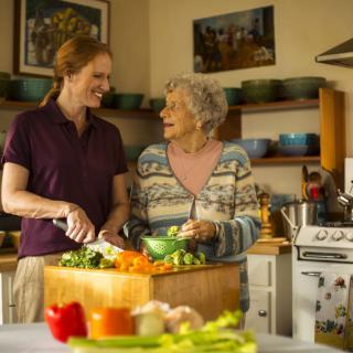 An elderly woman and a younger woman are smiling and chatting while preparing vegetables together in a cozy kitchen.