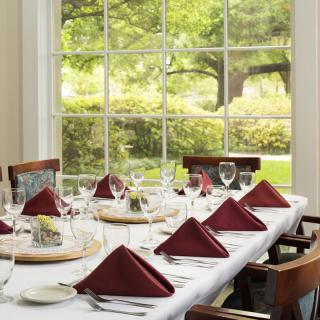 An elegantly set dining table with maroon napkins and wine glasses, bathed in natural light from a large window overlooking a garden.