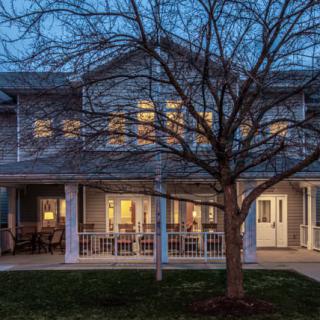 A cozy house with its lights on in the evening, featuring a front porch and tree in the yard.