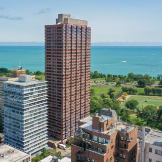A large skyscraper stands tall near a lake and park, surrounded by smaller buildings in an urban setting.