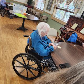 An elderly woman in a wheelchair engaging with a device at a communal table.