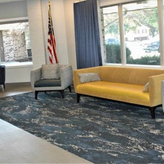 A modern lobby with a yellow sofa and a gray chair, adorned with a decorative rug and natural light from large windows.