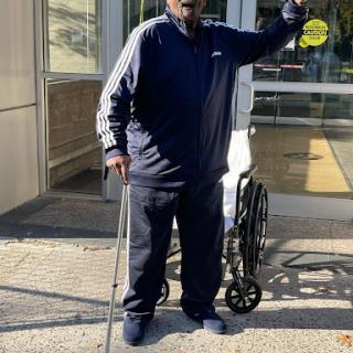An elderly man stands outdoors, gesturing while holding a walking stick next to a wheelchair.