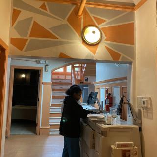 A person stands at a reception desk with abstract wall art and a clock above.