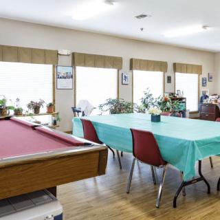 A recreation room featuring a billiards table and a dining area with tables and chairs, surrounded by windows and plants.