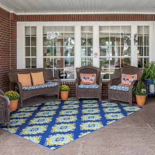 A cozy patio setup with wicker chairs, colorful cushions, a patterned rug, and potted plants against a brick wall.