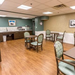 A clean, well-lit dining area with tables and chairs, featuring a kitchenette in the background.