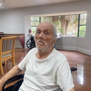 An elderly man seated in a care facility, with another resident in a wheelchair in the background.