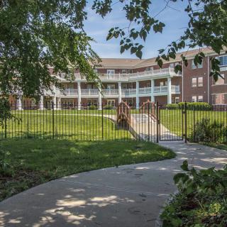 A pathway leads to a brick building with white columns, surrounded by greenery and enclosed by a fence.