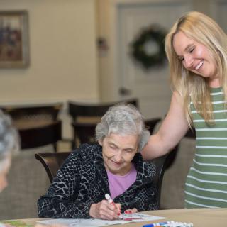 A young woman smiles as she interacts with an elderly woman engaged in an art activity.