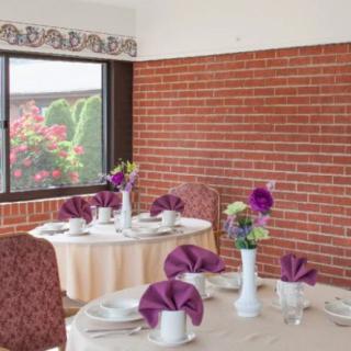 A cozy dining area with tables elegantly set with purple napkins and floral centerpieces, against a backdrop of a brick wall and window view of greenery.
