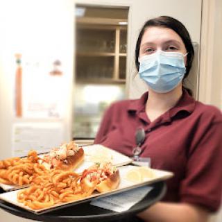 A staff member in a mask serving hot dogs and fries on a tray.
