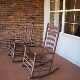 Two wooden rocking chairs on a porch with a brick wall and window.