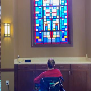 A person in a wheelchair sits in contemplation, facing a beautifully designed stained glass window that casts colorful light within the serene interior space.