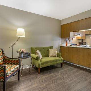 A cozy living space with a green sofa, patterned armchair, and a kitchenette in the background.