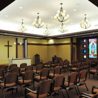 A chapel interior with rows of chairs, chandeliers, stained glass window, and a cross at the front.
