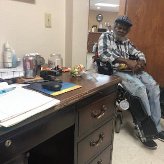A man seated in a wheelchair in an office setting, accompanied by a desk and various items.