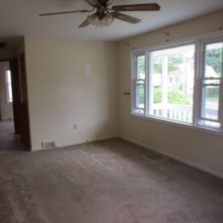An empty living room with a ceiling fan, carpeted floor, and large windows letting in natural light.