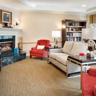 A cozy living room with a fireplace, featuring a sofa and armchairs, surrounded by a bookshelf and lamps.