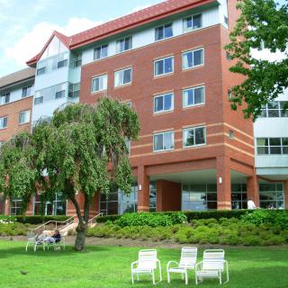 A multi-story brick residence building with chairs on the lawn and trees surrounding it.