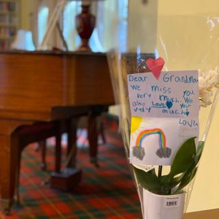 A flower bouquet with a handwritten note stands in front of a grand piano in a cozy room.