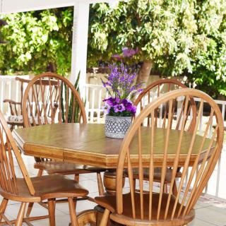 A cozy outdoor patio with a wooden table, surrounded by chairs and adorned with a vase of flowers in sunlight.