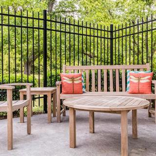 A cozy outdoor patio with wooden furniture and colorful cushions, surrounded by a black iron fence and greenery.