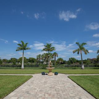 A tranquil landscape featuring a paved pathway leading to a fountain, surrounded by palm trees and a lush lawn under a clear blue sky.