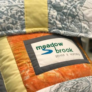 A close-up view of a colorful quilt featuring a label from 'Meadow Brook'.