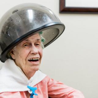 An elderly woman smiling while sitting under a hair dryer at a salon.