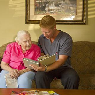 A caregiver and an elderly woman sit together on a couch, looking at a book.
