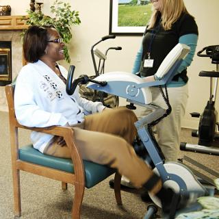 A person using exercise equipment with the assistance of a healthcare professional in a rehabilitation setting.
