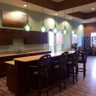 A modern kitchen interior with a large countertop and bar stools, illuminated by pendant lights.