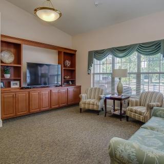 A cozy living room with a large television, sofa, and armchairs arranged around a coffee table, featuring large windows and elegant curtains.