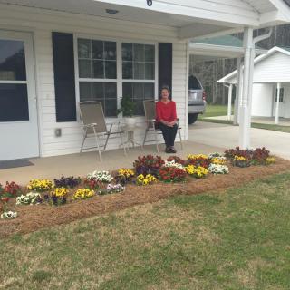 A person sitting on the porch of a house, surrounded by a garden with vibrant flowers.