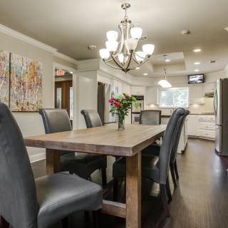 A modern dining room with a wooden table, elegant chairs, and a chandelier, opening into the kitchen area.