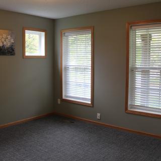 An empty room with carpet flooring, two large windows with blinds, and a small piece of wall art.
