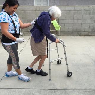 A caregiver assists an elderly person using a walker outside.