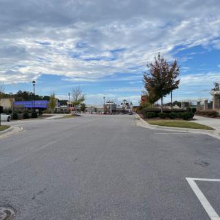 An empty parking lot under a cloudy sky with trees and buildings in the background.