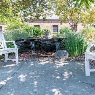 Tranquil garden setting with white chairs and a cascading water fountain under the shade of trees.