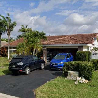A suburban house with a driveway, featuring two cars parked in front of a garage, surrounded by palm trees under a partly cloudy sky.