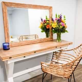 A stylish vanity setup with a large wooden-framed mirror, accompanied by a rustic chair and a vibrant bouquet of flowers in a vase.