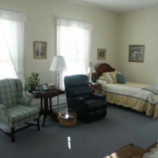 A cozy bedroom with a bed, two chairs, and natural light from the windows.