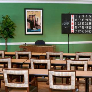 A classroom with rows of chairs and desks, green walls, a decorative plant, a framed poster, and a bingo board at the front.