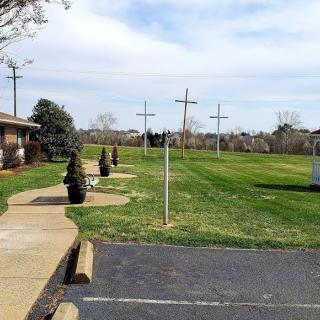 Three crosses stand on a grassy lawn next to a pathway and building, under a cloudy sky.