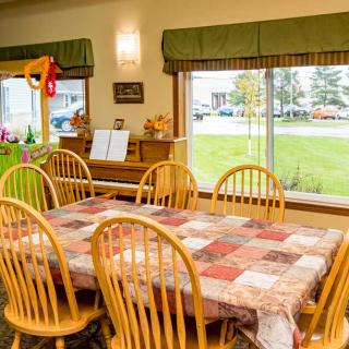 A cozy dining room featuring a large wooden table with a patterned tablecloth, surrounded by matching chairs. Large windows provide a view of the outside, and a piano is situated against the wall.