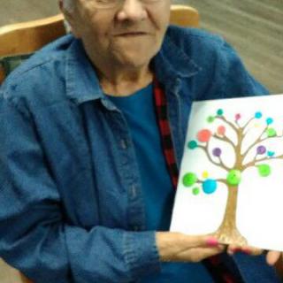 An elderly woman holding a colorful painting of a tree.