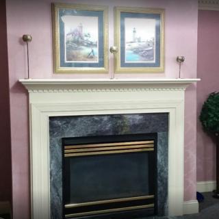 A cozy fireplace with a white mantel, adorned by dual framed artworks and set against a pink wall.