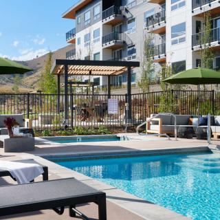 A modern apartment building features a luxurious outdoor swimming pool area with loungers and patio furniture on a sunny day.
